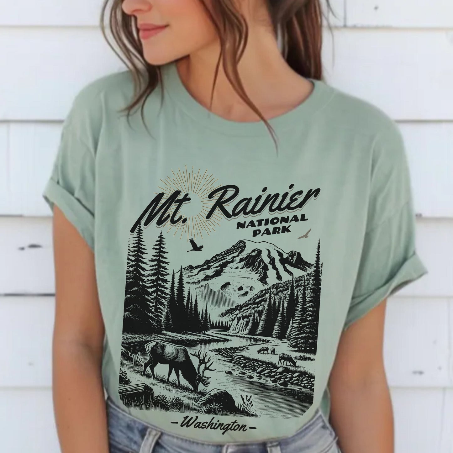A woman wearing a sage green graphic t-shirt featuring a black and white print of Mt. Rainier National Park, paired with light-wash denim jeans, set against a white wooden wall background.