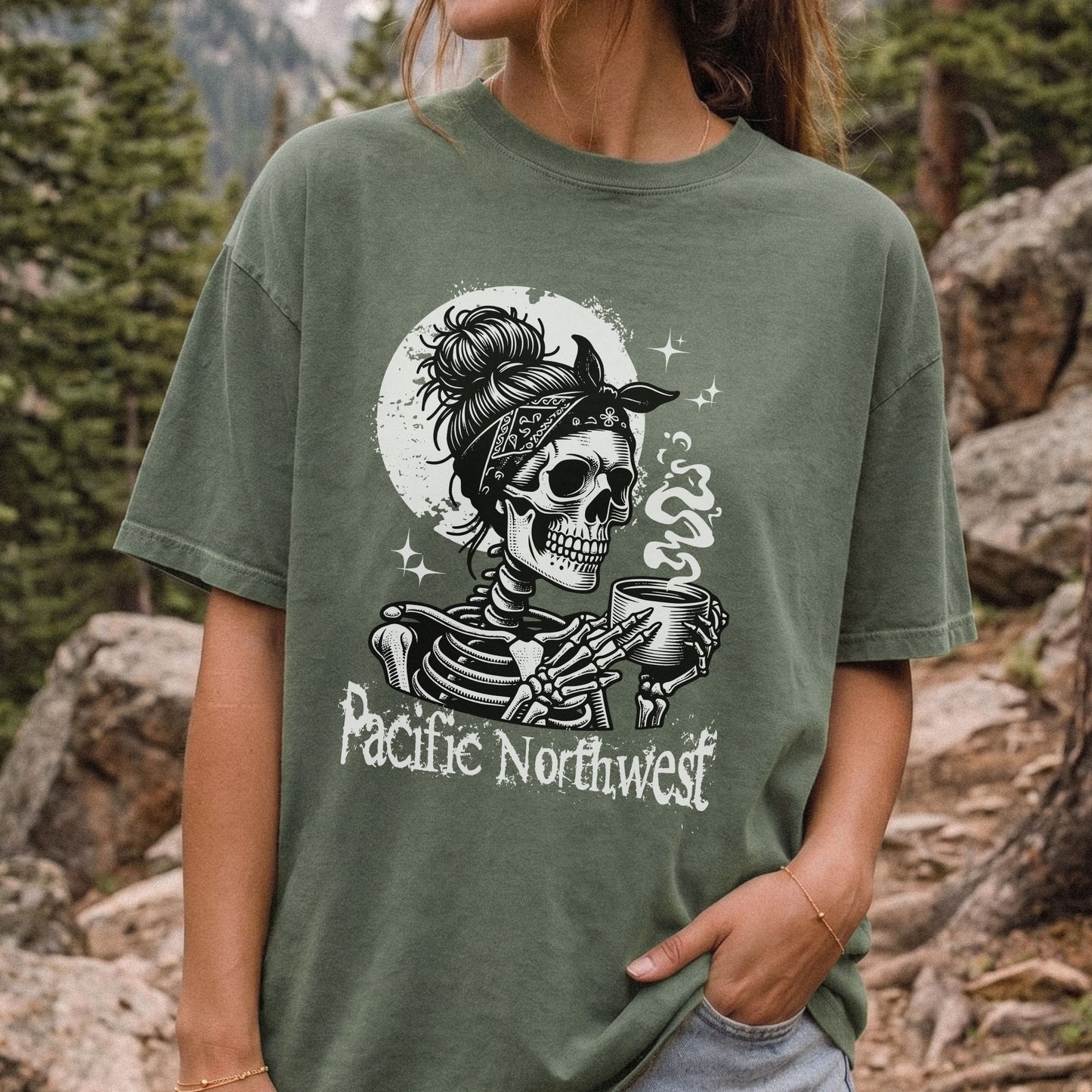 A woman wearing an oversized moss green graphic t-shirt in an outdoor mountain setting. The t-shirt features a black and white graphic of a skeleton wearing a bandana and holding a mug, with the text 'Pacific Northwest' printed below.