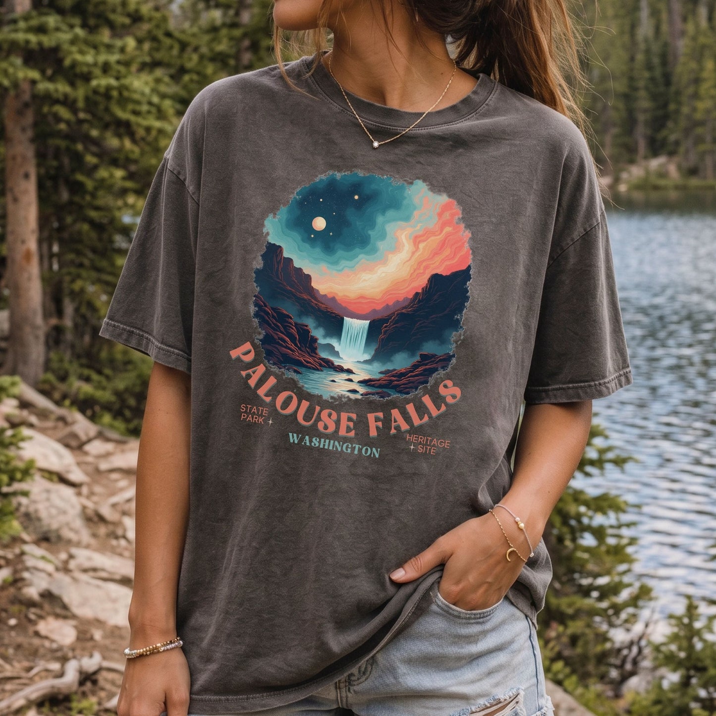A woman wearing a faded charcoal gray graphic t-shirt featuring a colorful illustration of a waterfall and stars, with the text 'PALOUSE FALLS WASHINGTON' printed below. She is standing outdoors near a lake and pine trees.