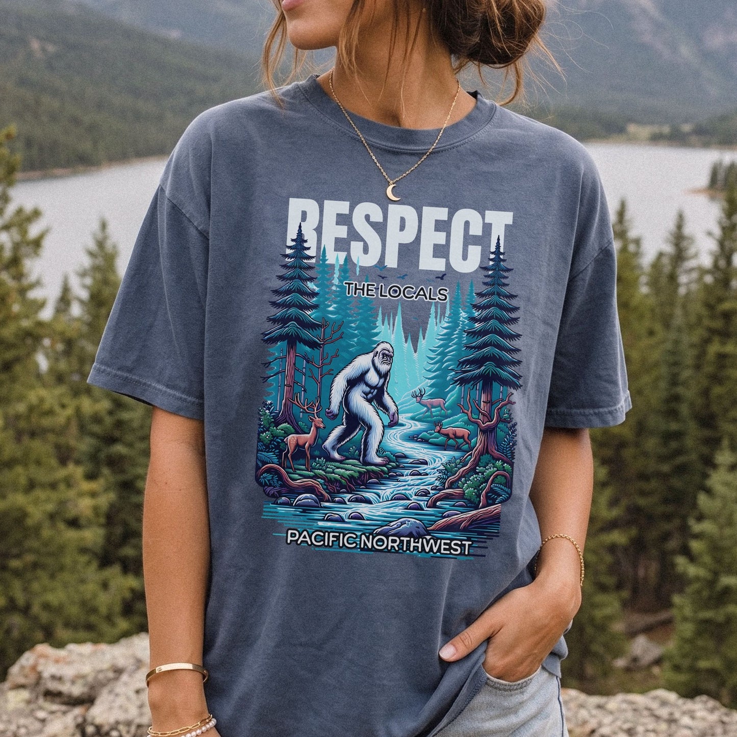 A woman wearing a faded blue graphic t-shirt with a 'Respect the Locals' Bigfoot design, set against a scenic outdoor background of a lake and pine trees.