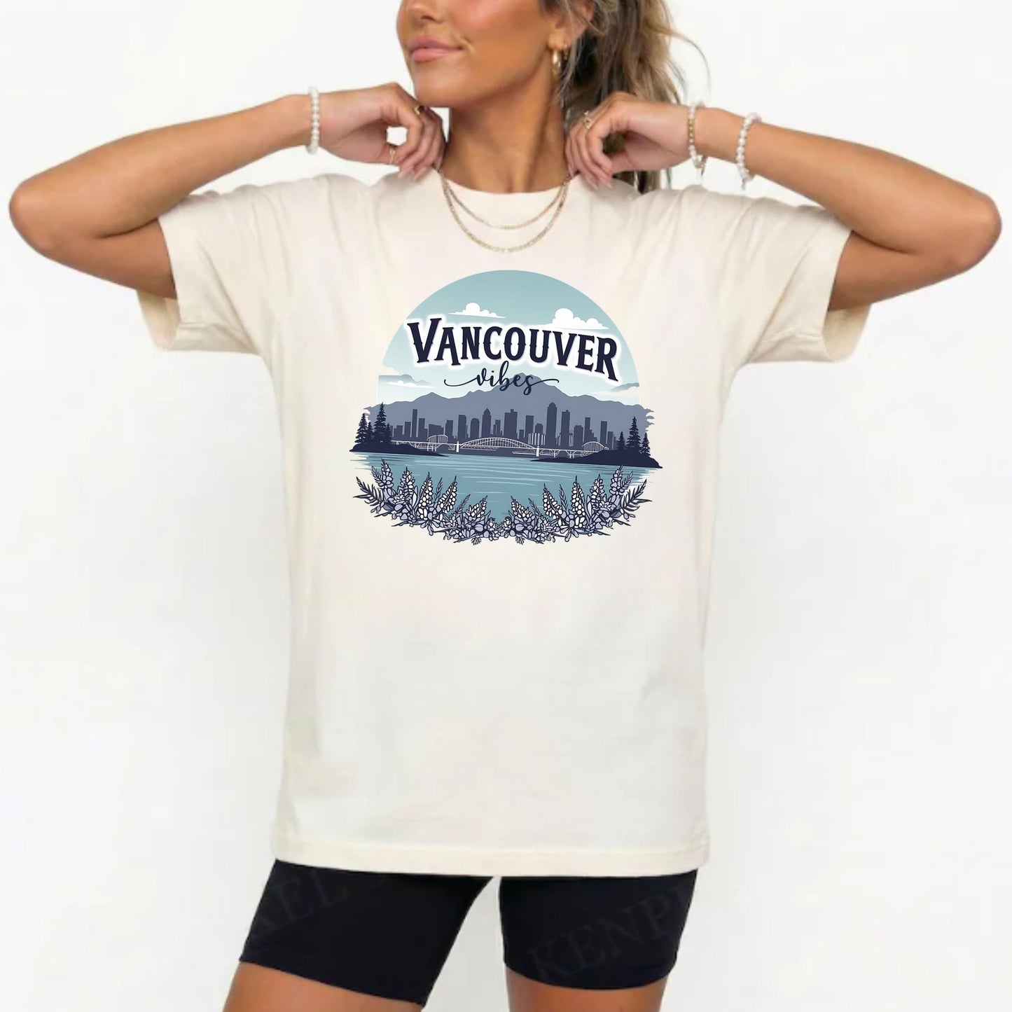 A woman wearing a cream-colored t-shirt with a graphic print of the Vancouver skyline and lake under a blue sky. She is also wearing black bike shorts and gold jewelry, posing against a plain white background.