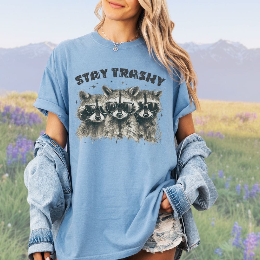 A person wearing a blue crew neck t-shirt with the phrase 'STAY TRASHY' printed above an illustration of three raccoons in sunglasses.