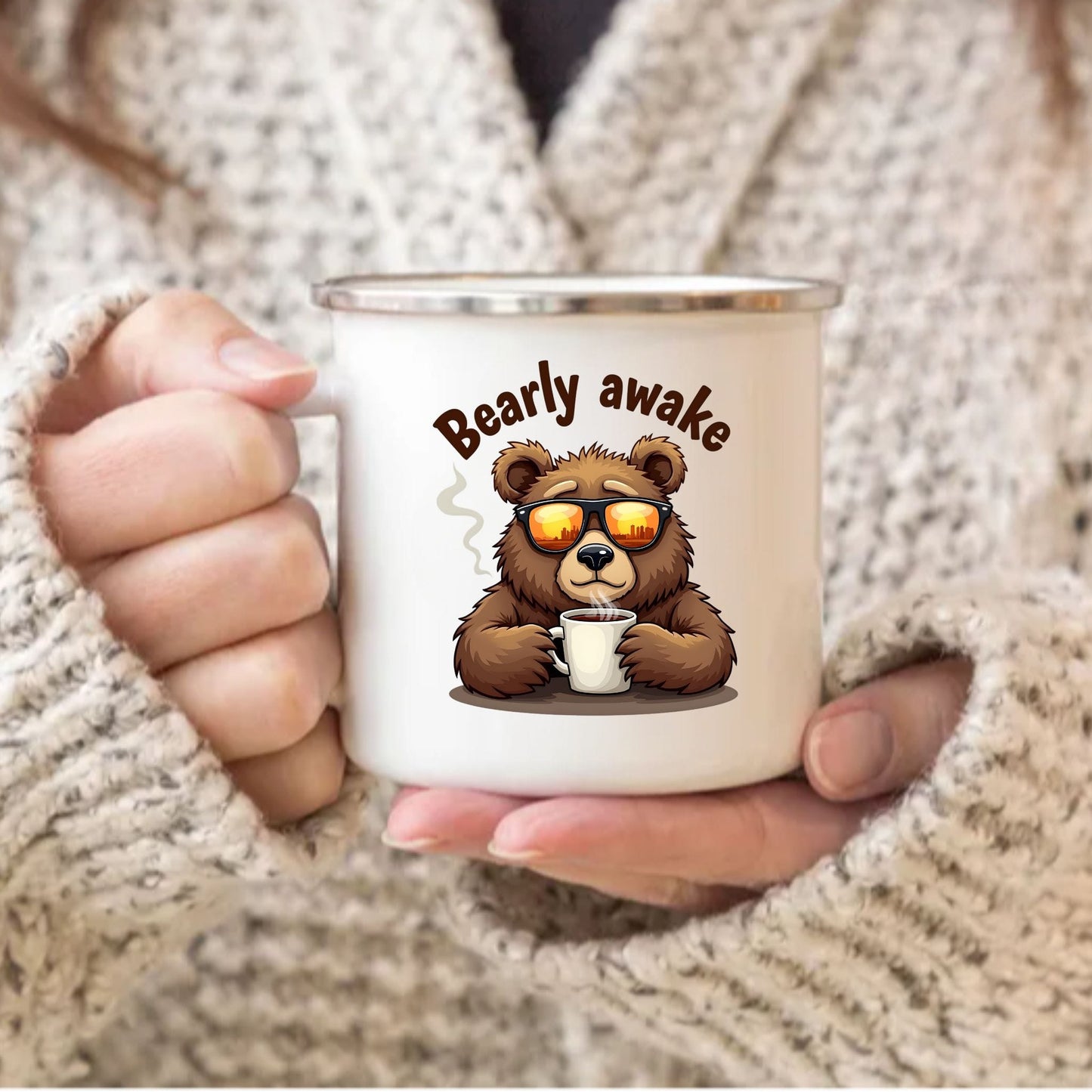 A person in a chunky cream knit sweater holds a white enamel-style mug with a cartoon brown bear wearing orange-tinted sunglasses and the words “Bearly awake” above it.