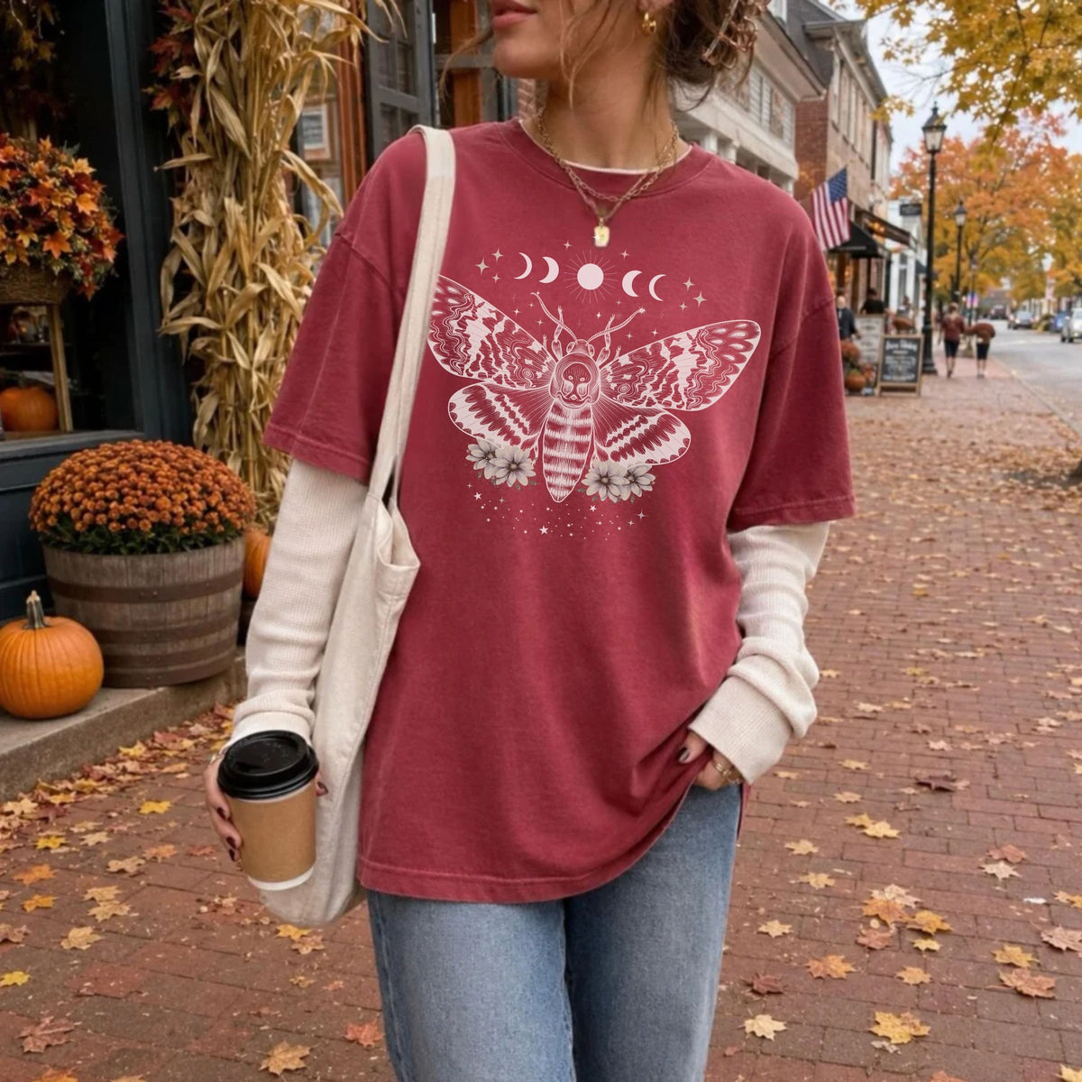 A woman wearing an oversized dusty rose graphic t-shirt with a white celestial moth and moon design, layered over a cream long-sleeve shirt, standing on a brick street in autumn.