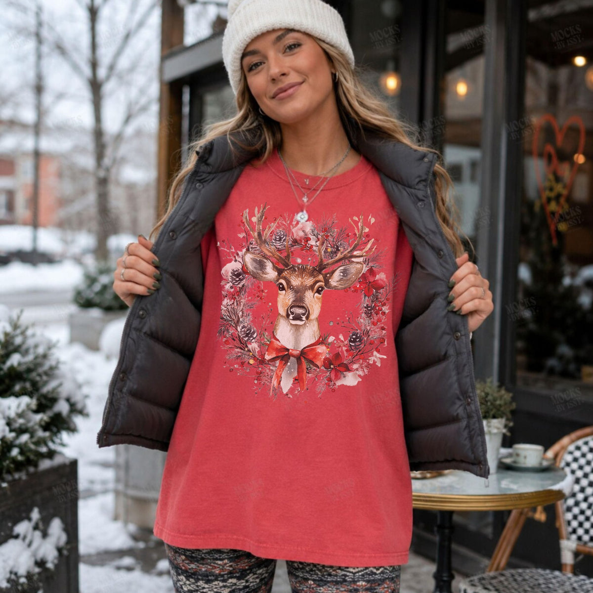 A woman wearing a red graphic t-shirt with a deer wreath design, layered under an open black puffer jacket, with a white knit beanie. She is standing outdoors in front of a storefront with snow visible on the ground and trees.