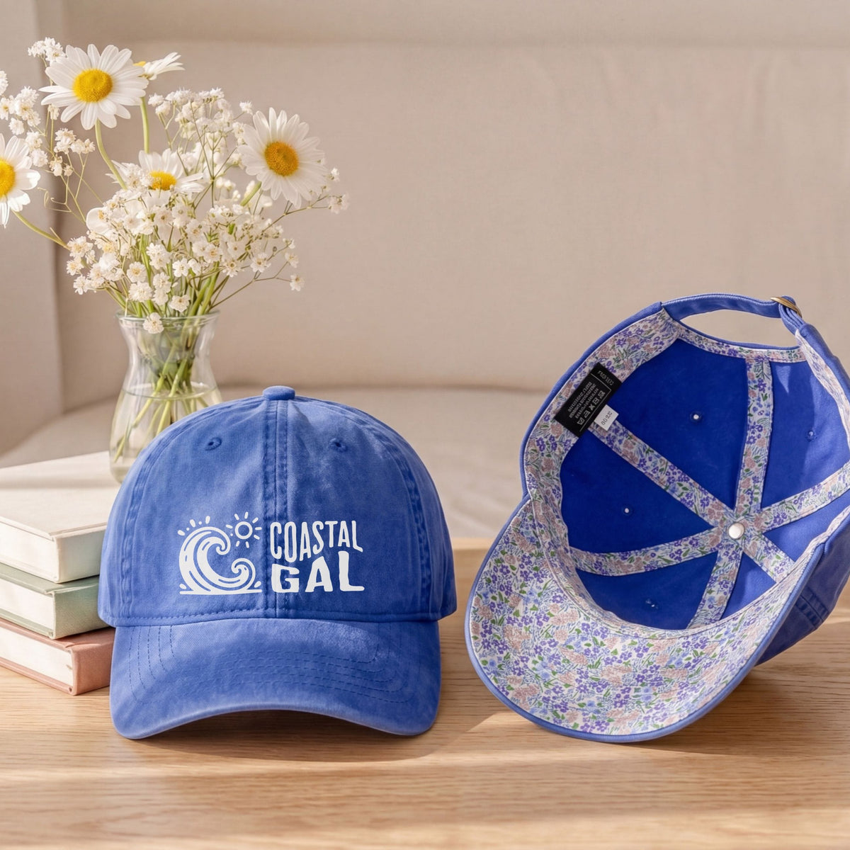 A blue baseball cap shown from the front and side on a wooden table, with a vase of white daisies and stacked books in the background. The front of the cap has a white 'COASTAL GAL' wave logo, and the inside of the cap features a glittery blue and silver pattern.