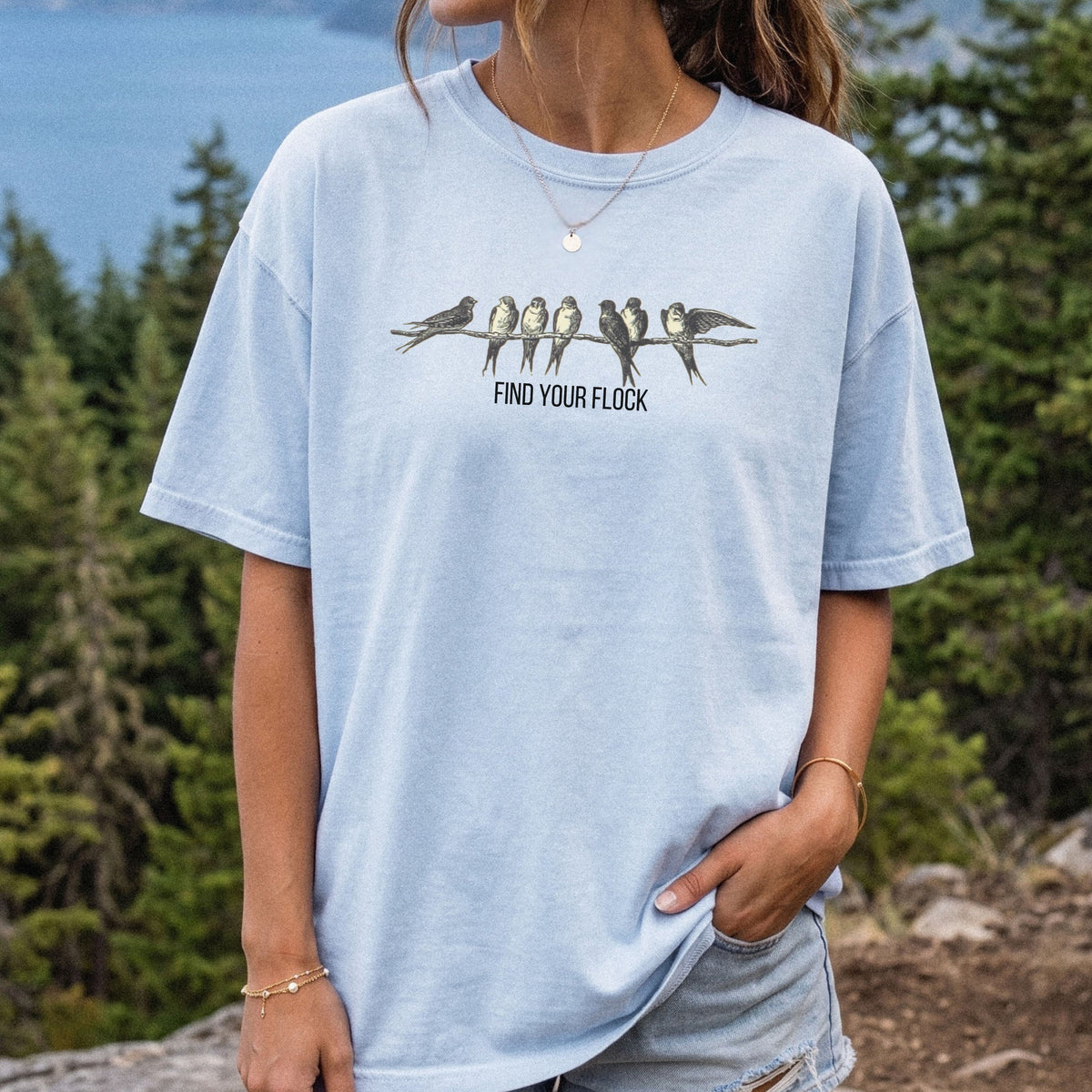 A woman wearing a light blue graphic t-shirt with a print of birds on a branch and the text 'FIND YOUR FLOCK' in front of a forest and lake background.
