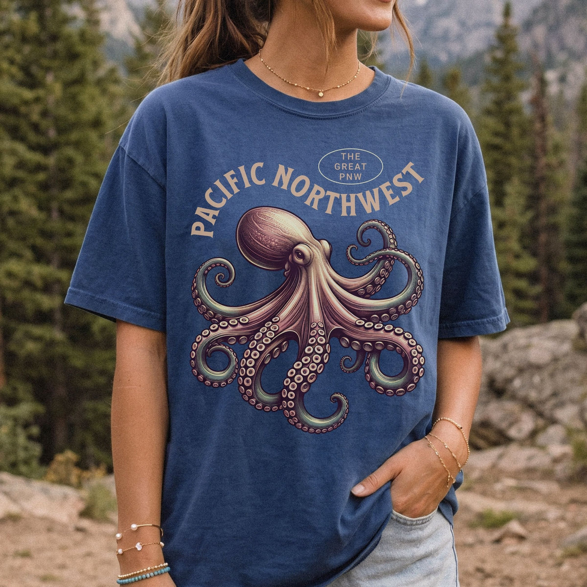A woman wearing a blue graphic t-shirt with a large octopus design and the text 'PACIFIC NORTHWEST' and 'THE GREAT PNW'. The background is a blurred outdoor forest scene.