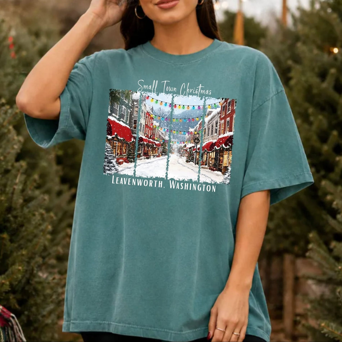 A woman wearing an oversized teal graphic t-shirt with a 'Small Town Christmas' print in Lewenworth, Washington, standing outdoors in front of Christmas trees.