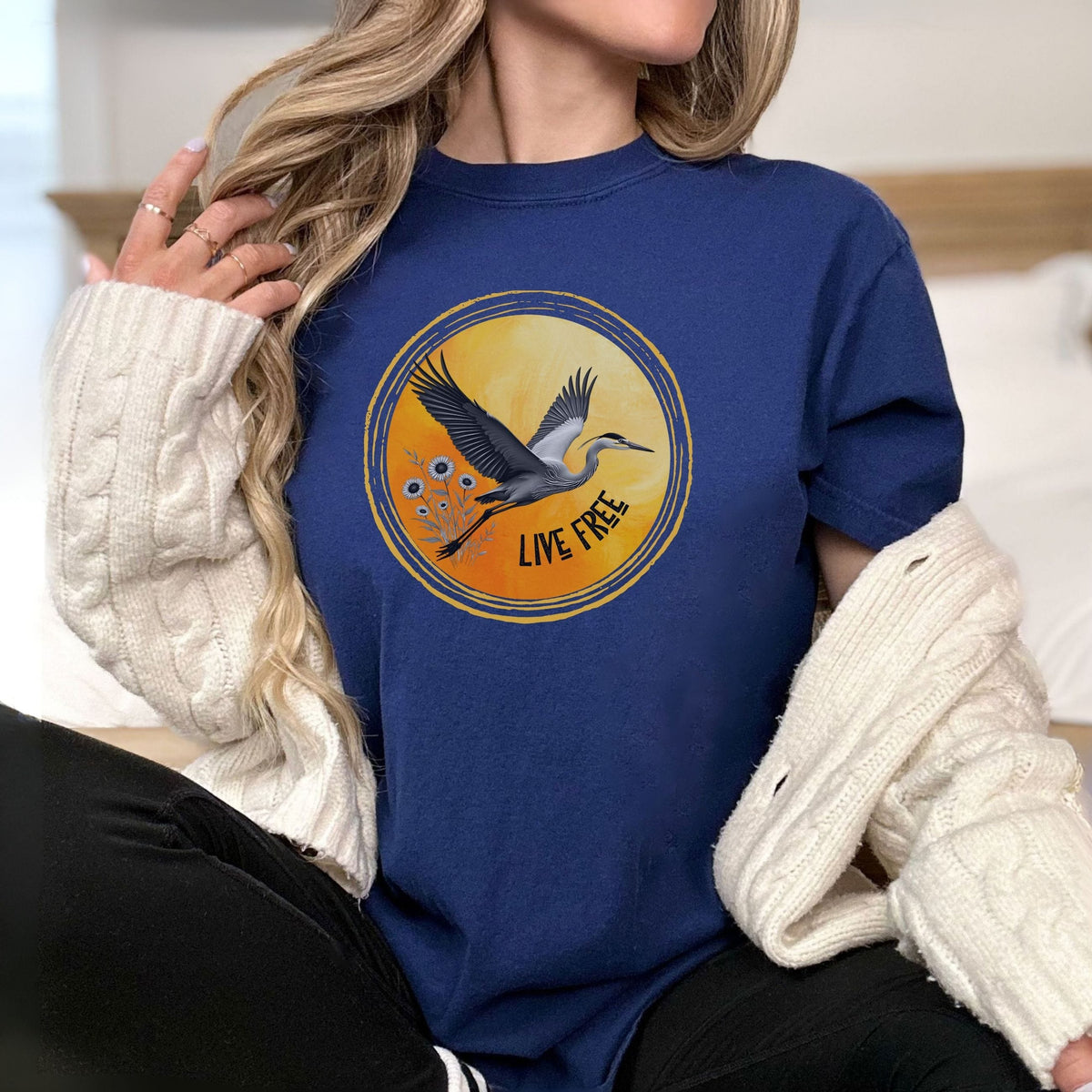 A woman wearing a navy blue crewneck t-shirt with a circular graphic of a heron and the words 'LIVE FREE'. She has a cream cable-knit sweater draped off her shoulders and is wearing black pants, set against a soft-focus indoor background.