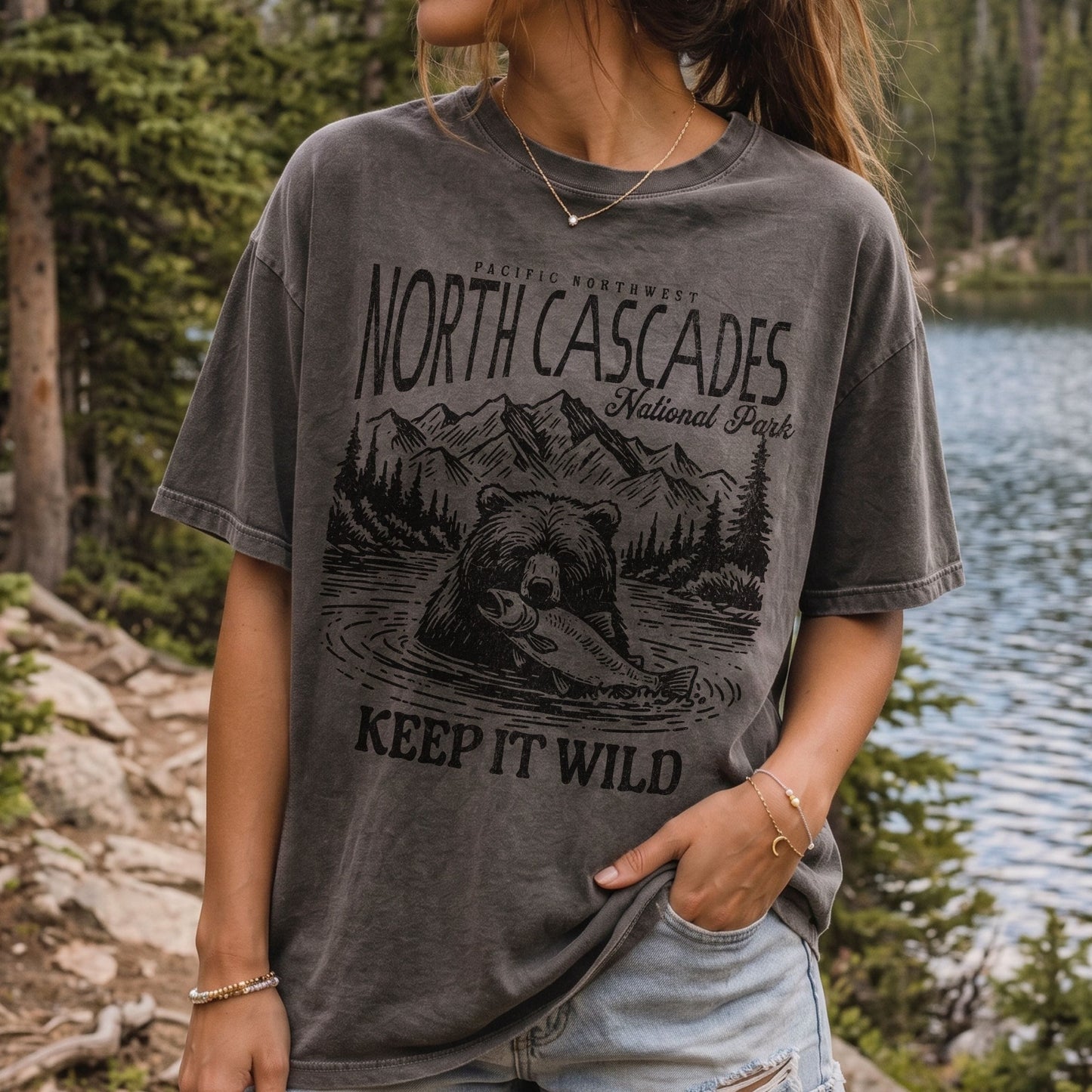 A woman wearing a faded dark gray graphic t-shirt with a black print of a bear, mountains, and the text 'NORTH CASCADES National Park' and 'KEEP IT WILD', set against a natural forest and lake background.