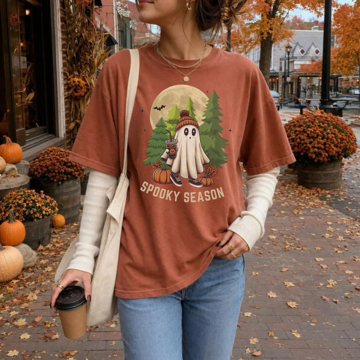 A woman wearing a rust-orange graphic t-shirt with a 'Spooky Season' cartoon ghost design, layered over a cream long-sleeve shirt and blue jeans, carrying a white tote bag in an autumn street setting.