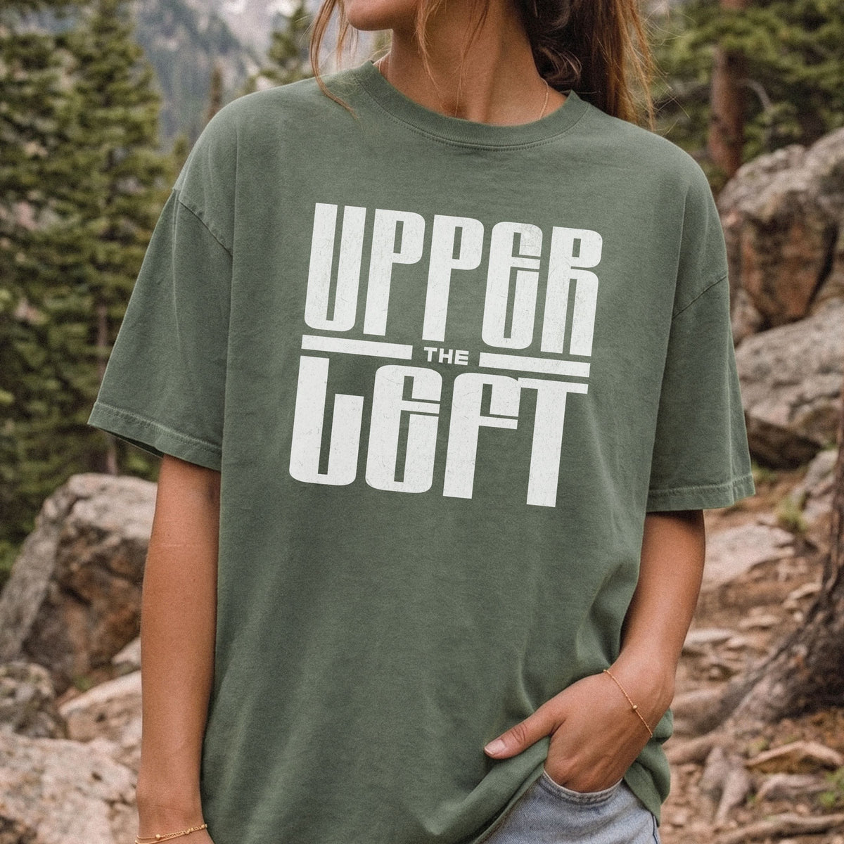 A woman wearing an oversized moss green t-shirt with a bold white graphic print that reads 'UPPER LEFT' against a natural outdoor background of trees and rocks.