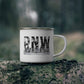 A white enamel camping mug with a silver metal rim and handle, featuring a black 'PNW' forest graphic, resting on a mossy log with a blurred forest background.