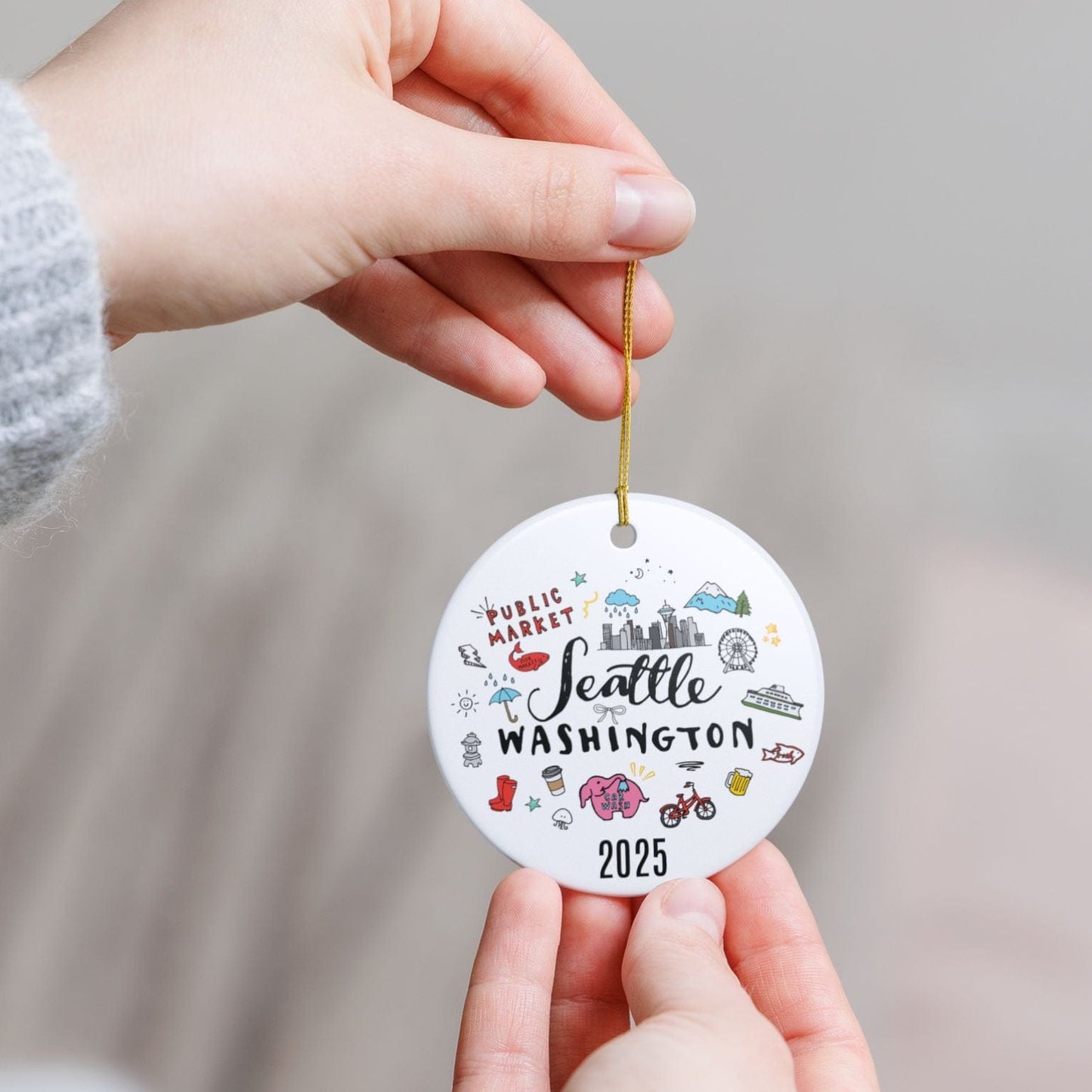 Hand holding a round ornament with 'Seattle, Washington 2025' text and illustrations.