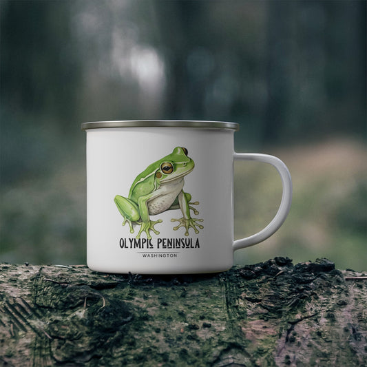 White enamel camp mug with a green frog illustration and Olympic Peninsula, Washington lettering, displayed outdoors on a mossy log.