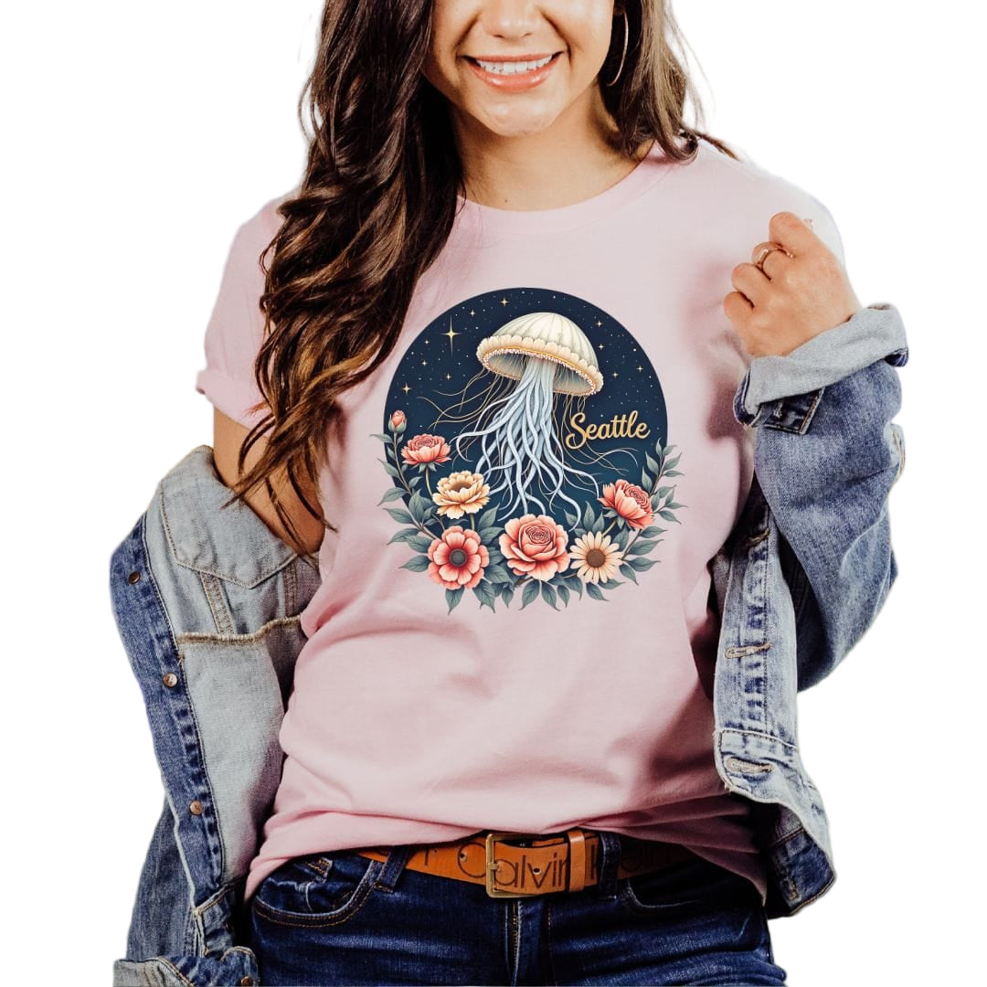 A woman wearing a pink t-shirt with a floral jellyfish design and the text 'Seattle' on it, standing in a field with mountains in the background.