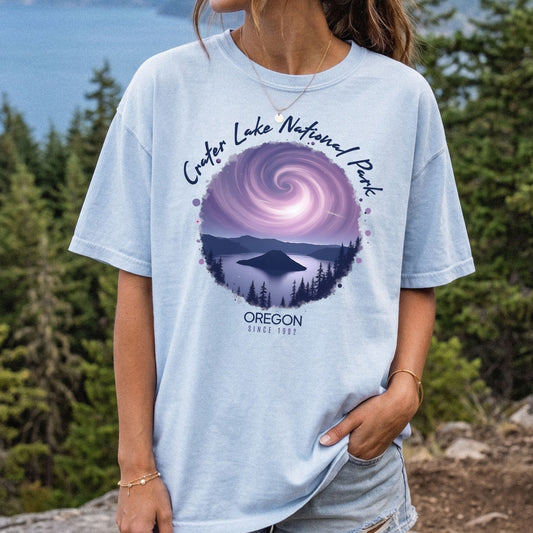 A woman wearing a Comfort Colors Chambray t-shirt with a vintage-style circular graphic of Crater Lake at night under a purple spiral galaxy, framed by white cursive text that reads Crater Lake National Park Oregon.
