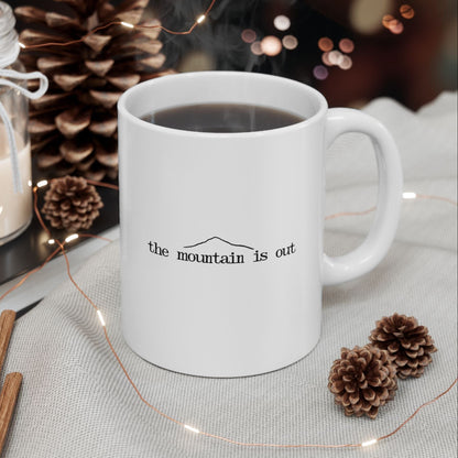 White mug with 'the mountain is out' text on a table with decorative items like a candle, oranges, and pine cones.