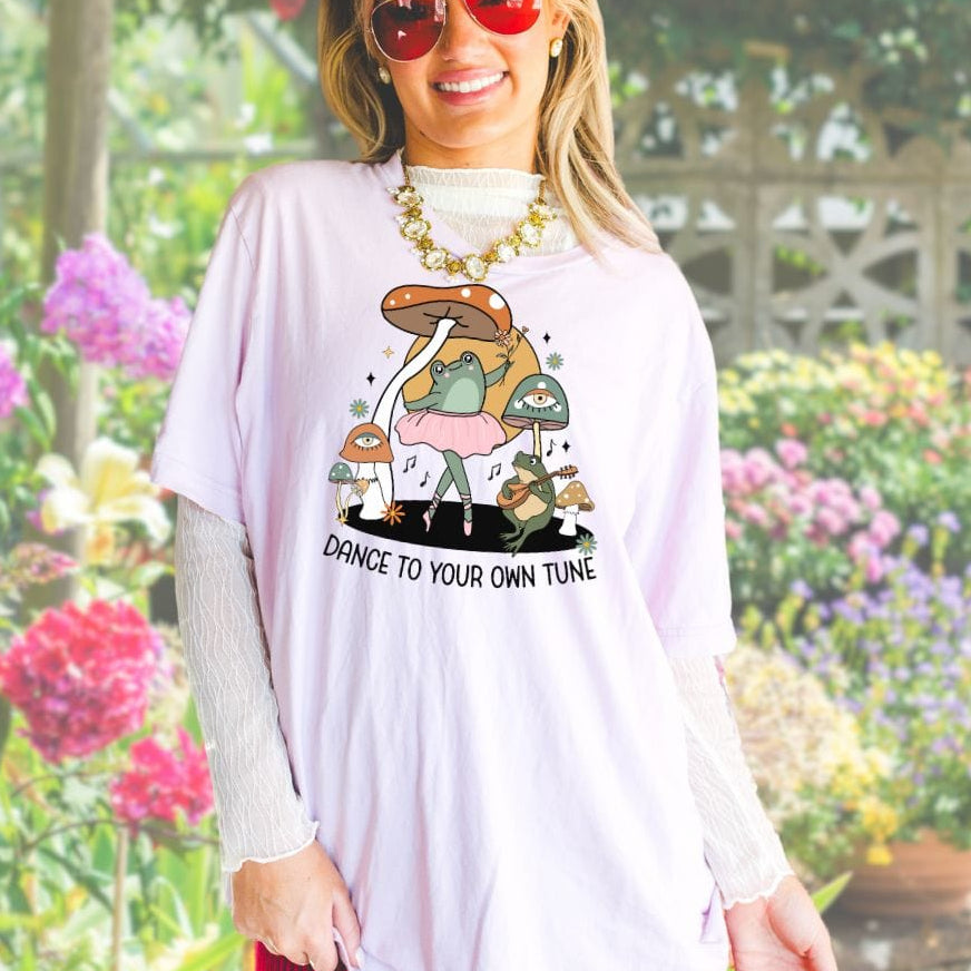 A woman wearing sunglasses and a pink t-shirt with a playful frog design and the text 'Dance to your own tune' on it, standing in a garden.