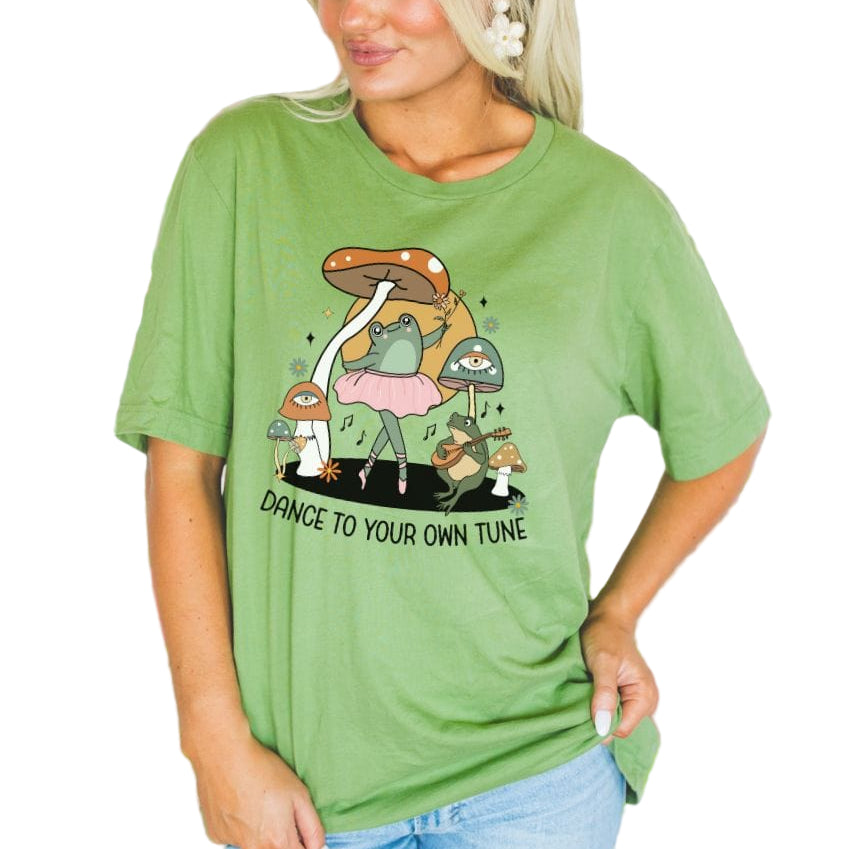 A woman wearing a green softstyle tee with a playful frog design and the text 'Dance to your own tune' on it.