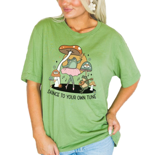 A woman wearing a green softstyle tee with a playful frog design and the text 'Dance to your own tune' on it.