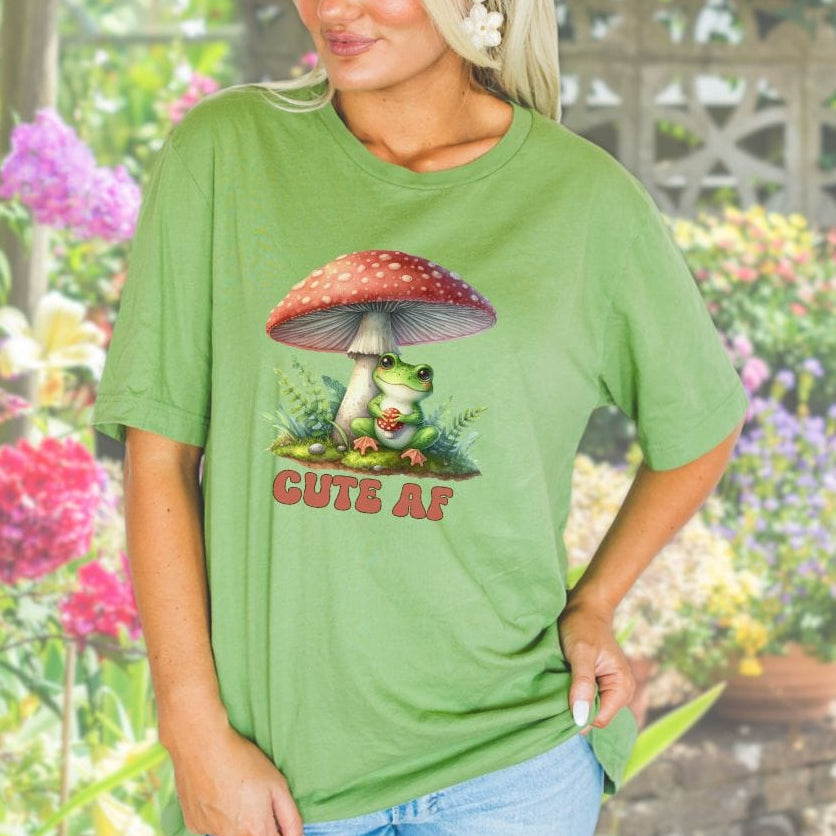 A woman wearing a green t-shirt with a frog and mushroom design, standing in a garden.