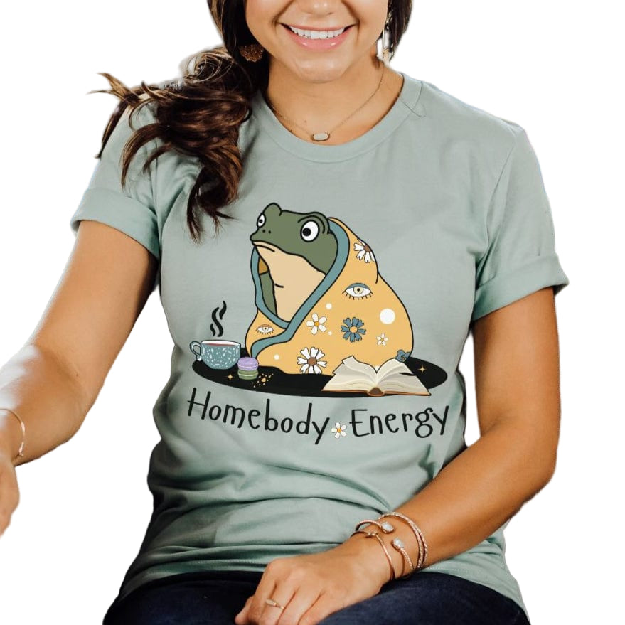 A person wearing a dusty blue t-shirt with a graphic of a frog and the text 'Homebody Energy'.