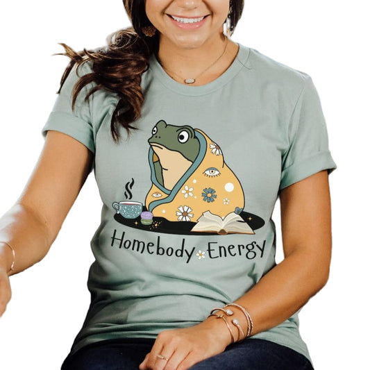 A person wearing a dusty blue t-shirt with a graphic of a frog and the text 'Homebody Energy'.