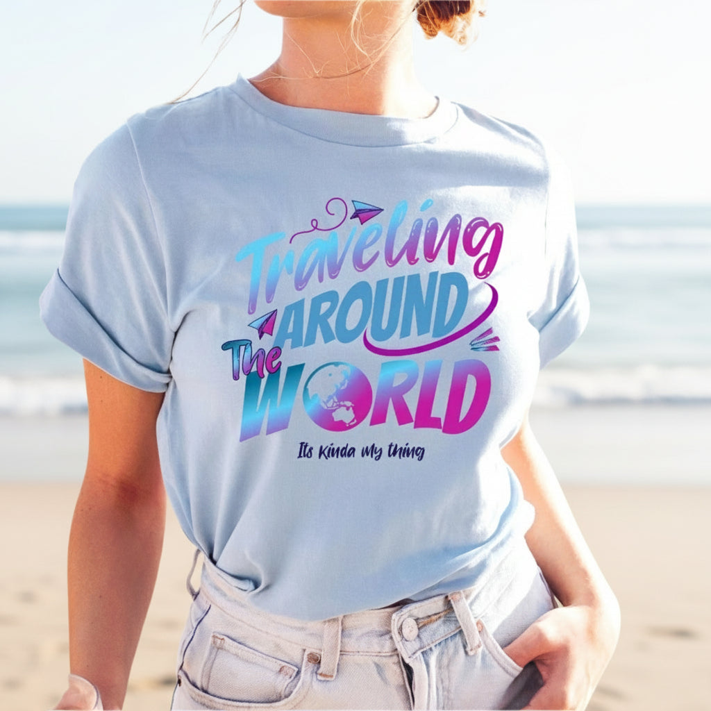 A woman wearing a light blue T-shirt with the text 'Traveling Around the World' printed on it, standing by the beach.