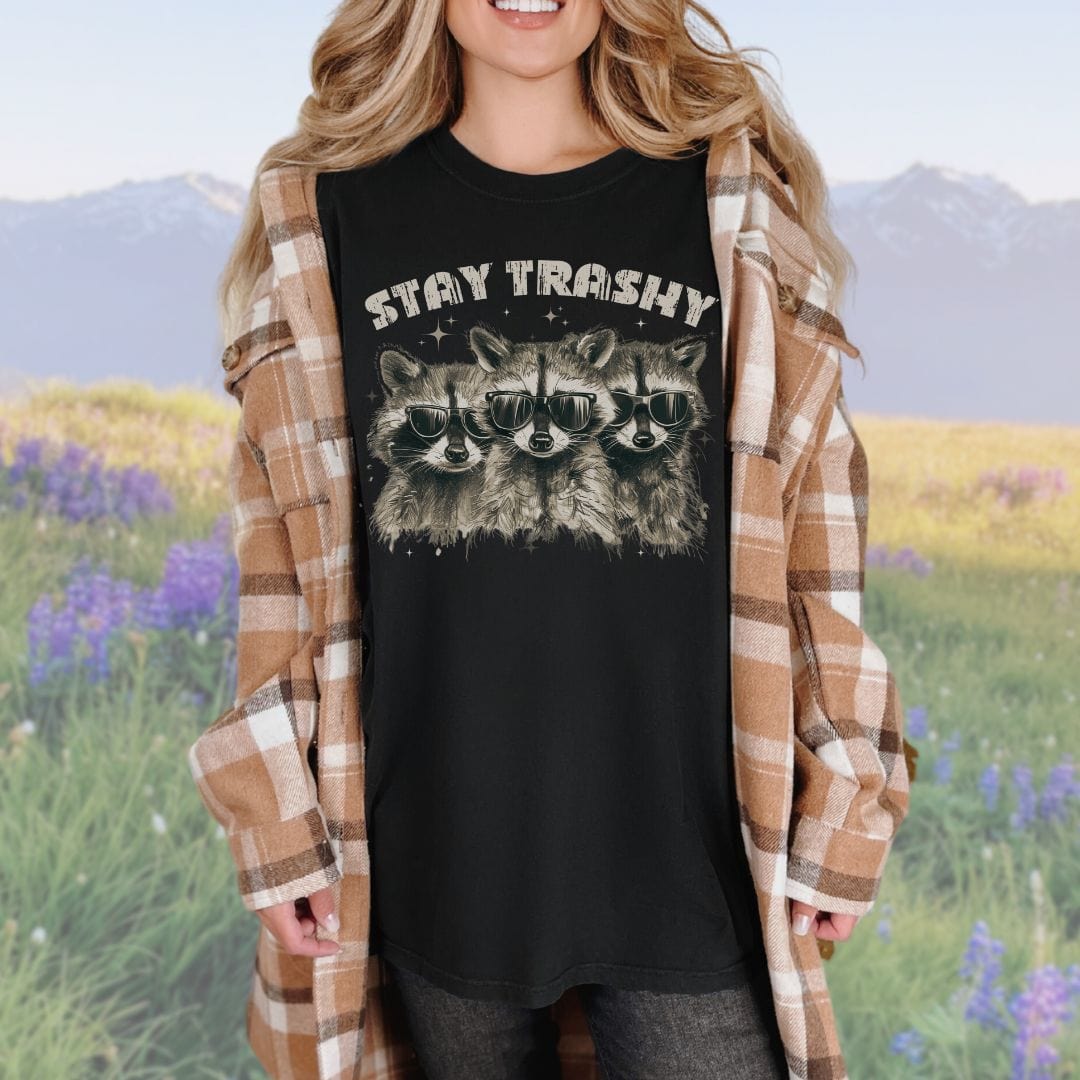 A person wearing a black crew neck t-shirt with the phrase 'STAY TRASHY' printed above an illustration of three raccoons in sunglasses.