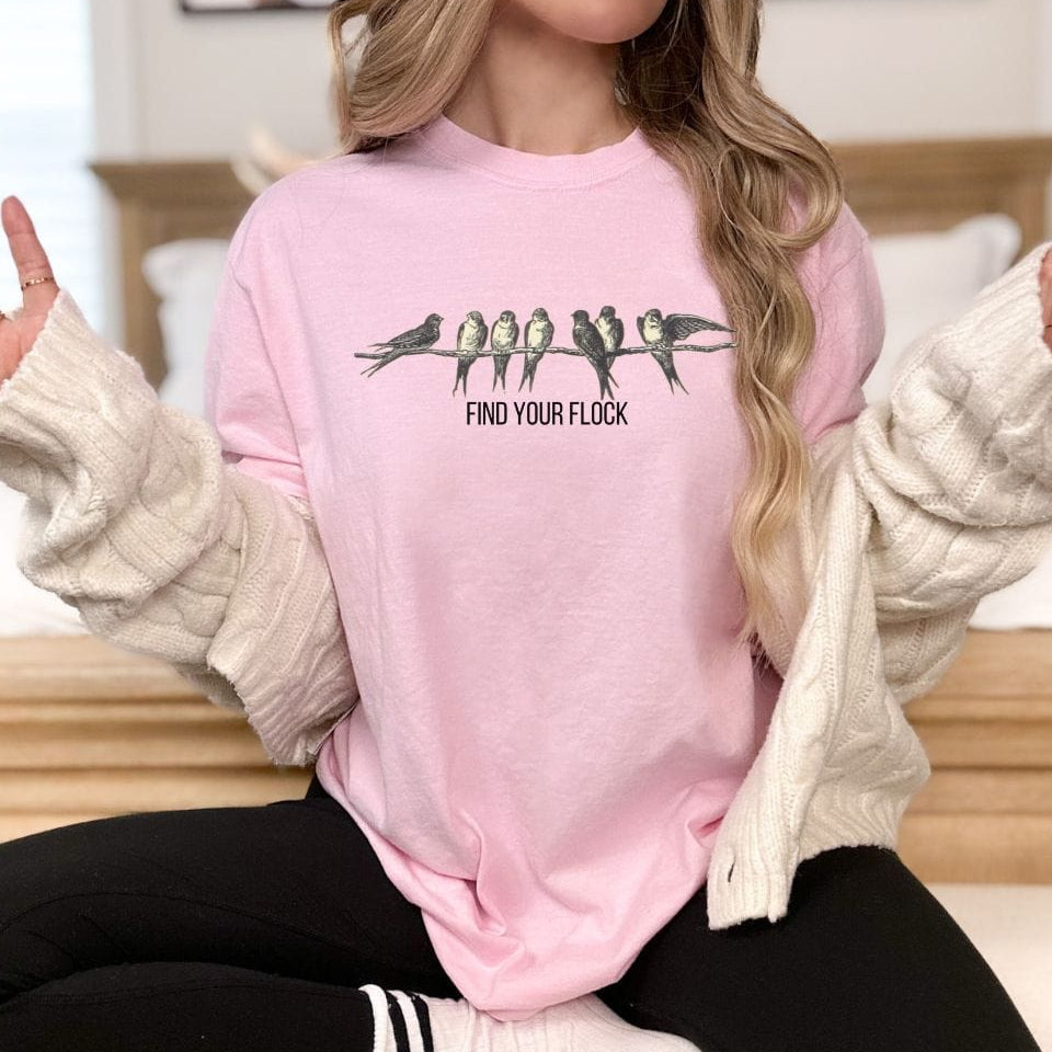 A person wearing a vintage-style pink shirt with bird illustrations and the text 'FIND YOUR FLOCK' on it, sitting in a cozy room with a knit blanket and a cup of coffee.