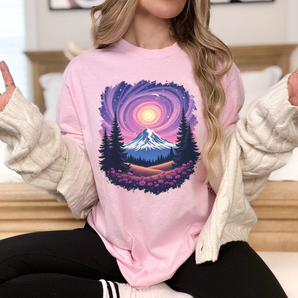 A person sitting and modeling a pink t-shirt with a vintage-style mountain and starry sky design.
