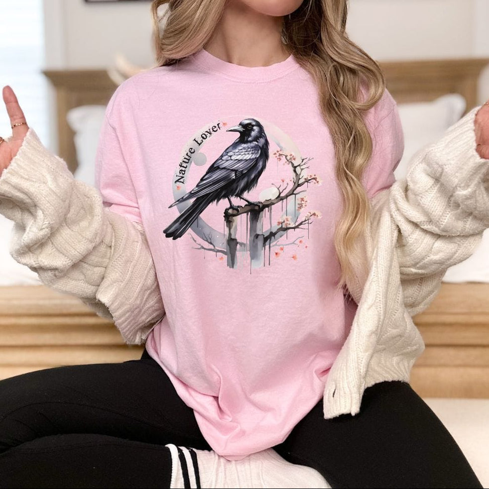 A person wearing a pink vintage-style t-shirt with a crow design and the text 'Nature Lover' on it, sitting on a white chair with denim shorts and a necklace.