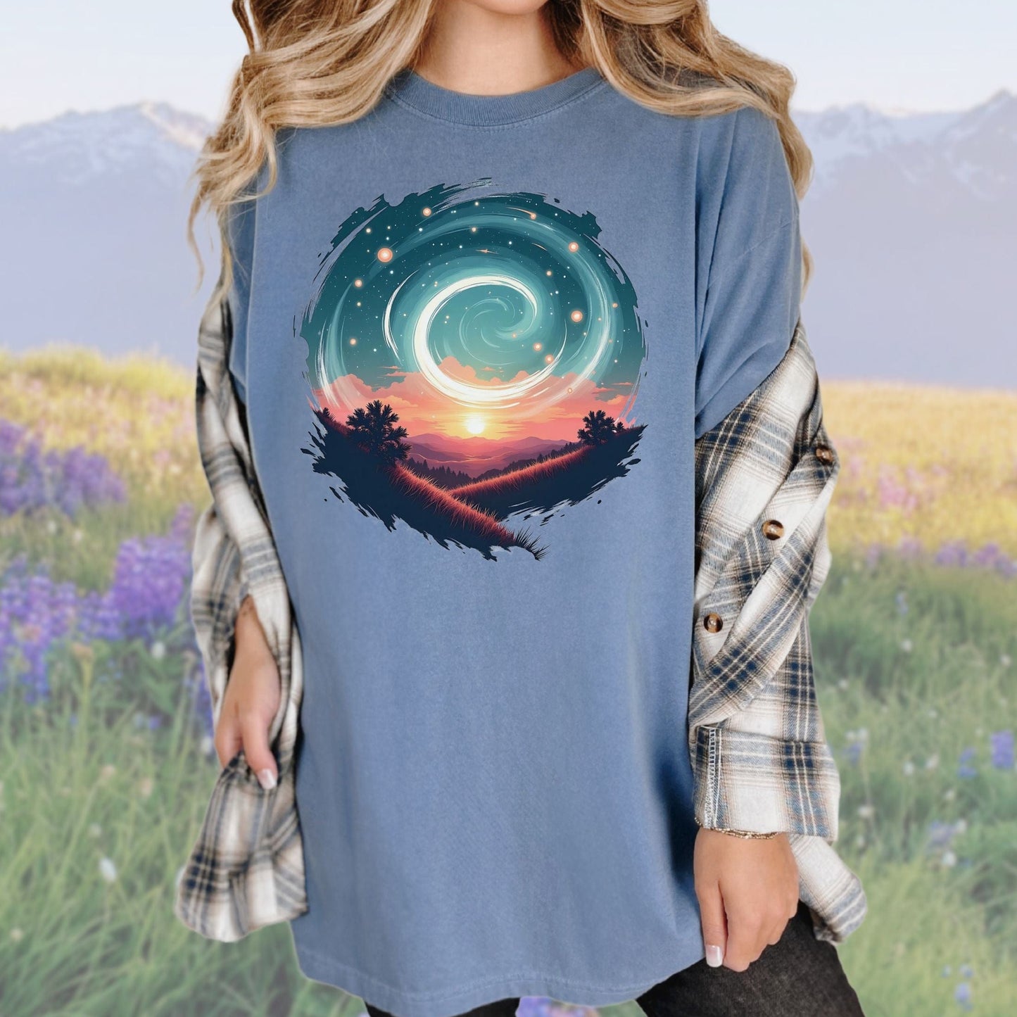 A person wearing a vintage-style graphic tee in a blue color with a swirl pattern in the design, standing in a field with mountains in the background.