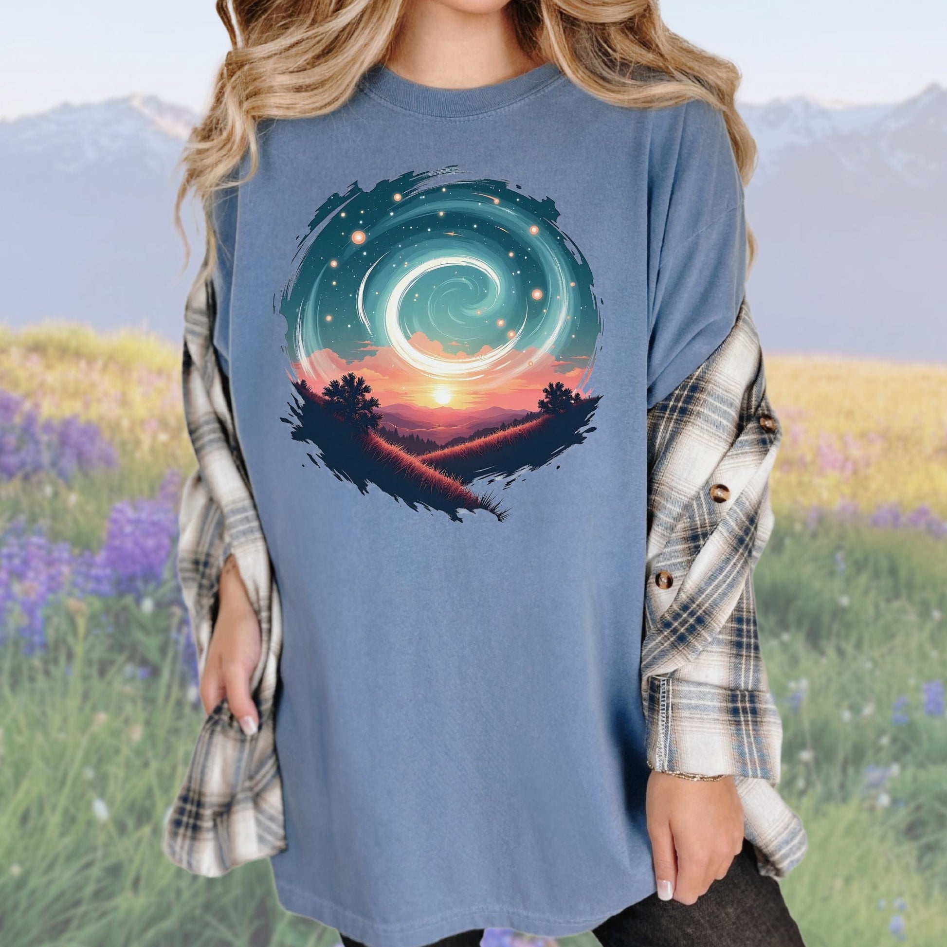 A person wearing a vintage-style graphic tee in a blue color with a swirl pattern in the design, standing in a field with mountains in the background.