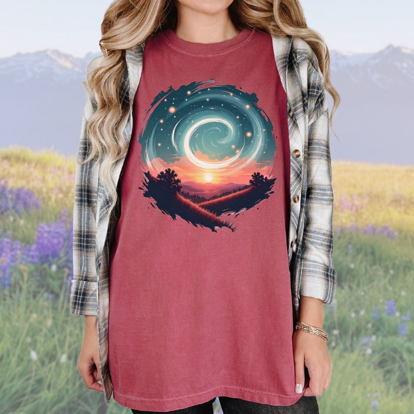 A person wearing a vintage-style graphic tee in a brick color with a swirl pattern in the design, standing in a field with mountains in the background.