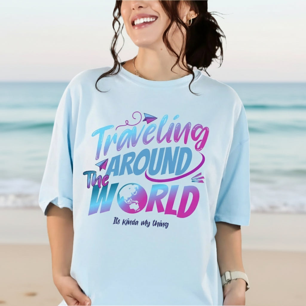 A person standing by the beach wearing a blue t-shirt with the text 'Traveling Around The World' in a colorful, vintage-style font.