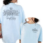 A person wearing a light blue t-shirt with floral line art design representing Skagit Valley, Washington. The shirt has a pocket and a back design with the text 'WALK AMONG THE FLOWERS'.