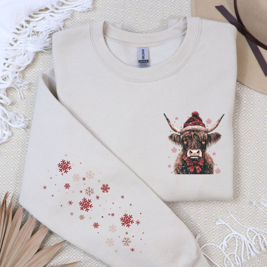 A folded cream sweatshirt laid flat on a patterned fabric background. The sweatshirt has a printed illustration of a highland cow wearing a red Santa hat and bow tie on the right chest, and scattered red and beige snowflakes on the left sleeve.
