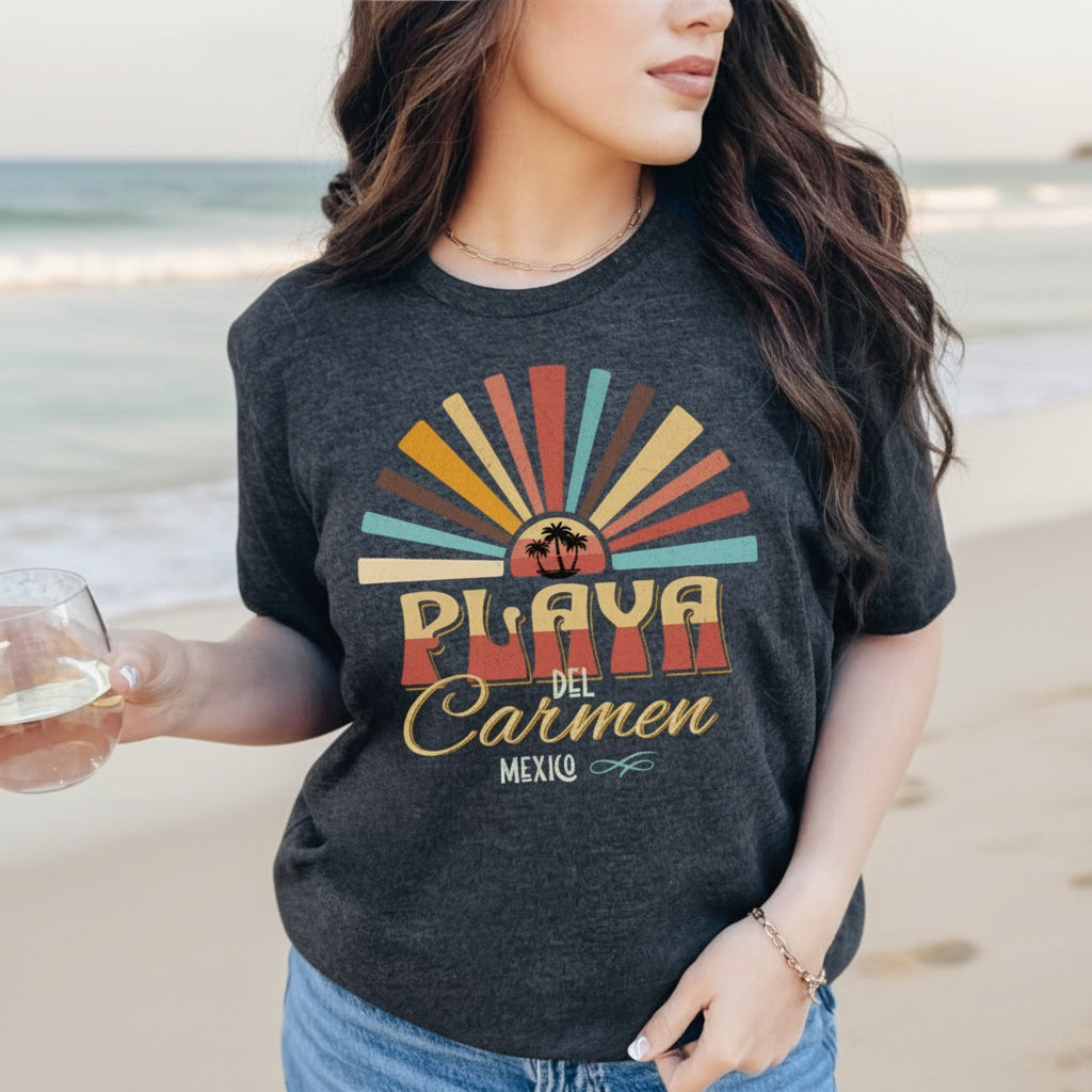 A person sitting by the beach wearing a dark gray t-shirt with 'Playa del Carmen Mexico' printed in a colorful text design.