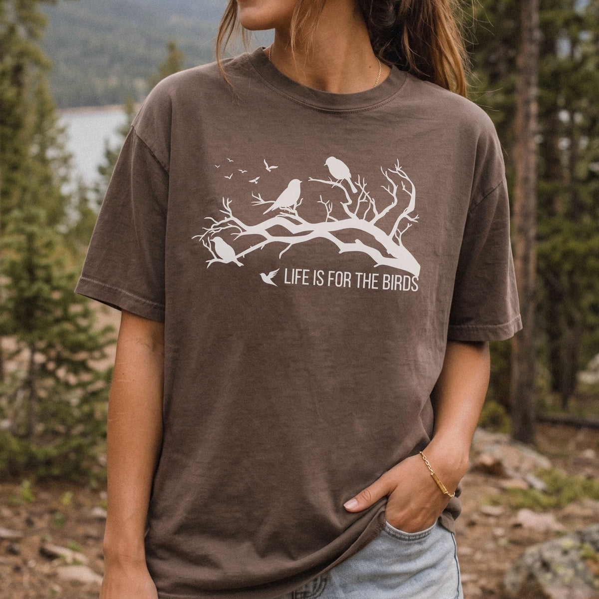 A woman wearing a brown t-shirt with a white graphic of birds on a branch and the text 'LIFE IS FOR THE BIRDS', photographed outdoors in a forest setting.
