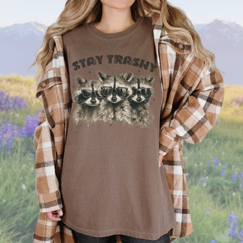 A person wearing a brown
 crew neck t-shirt with the phrase 'STAY TRASHY' printed above an illustration of three raccoons in sunglasses.