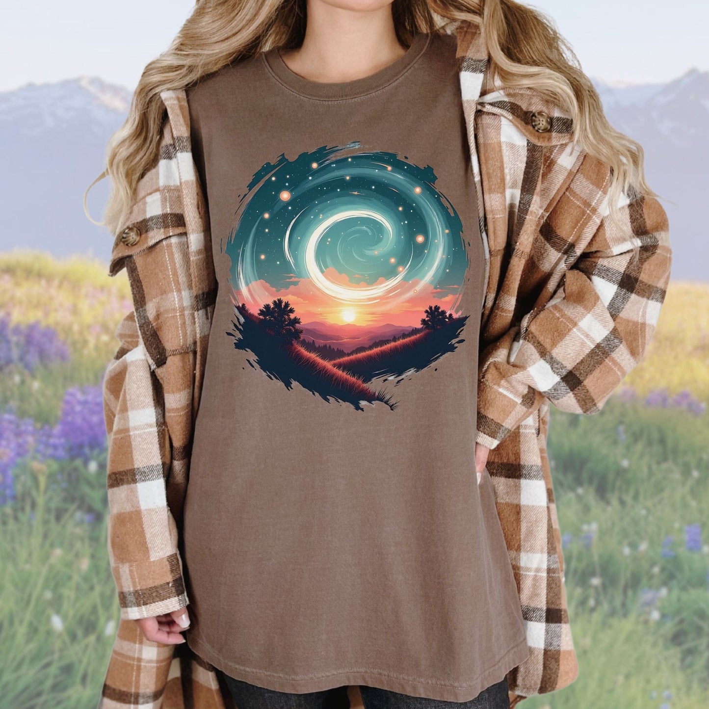 A person wearing a vintage-style graphic tee in an espresso color with a swirl pattern in the design, standing in a field with mountains in the background.