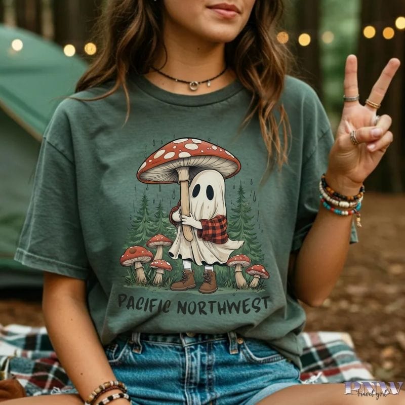 Person wearing a moss colored t-shirt with a ghost and mushroom design in a forest setting