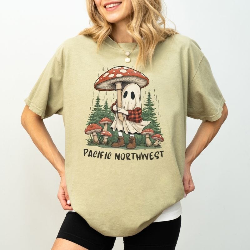 Woman wearing a sage green Comfort Colors t-shirt with an illustration of a cute ghost holding a mushroom umbrella in a rainy Pacific Northwest forest.
