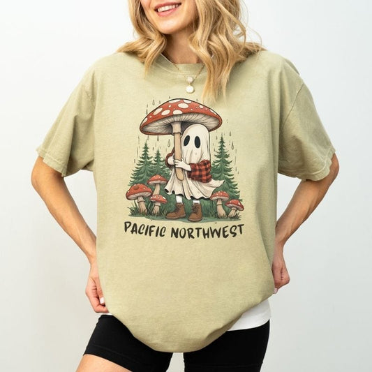 Woman wearing a sage green Comfort Colors t-shirt with an illustration of a cute ghost holding a mushroom umbrella in a rainy Pacific Northwest forest.