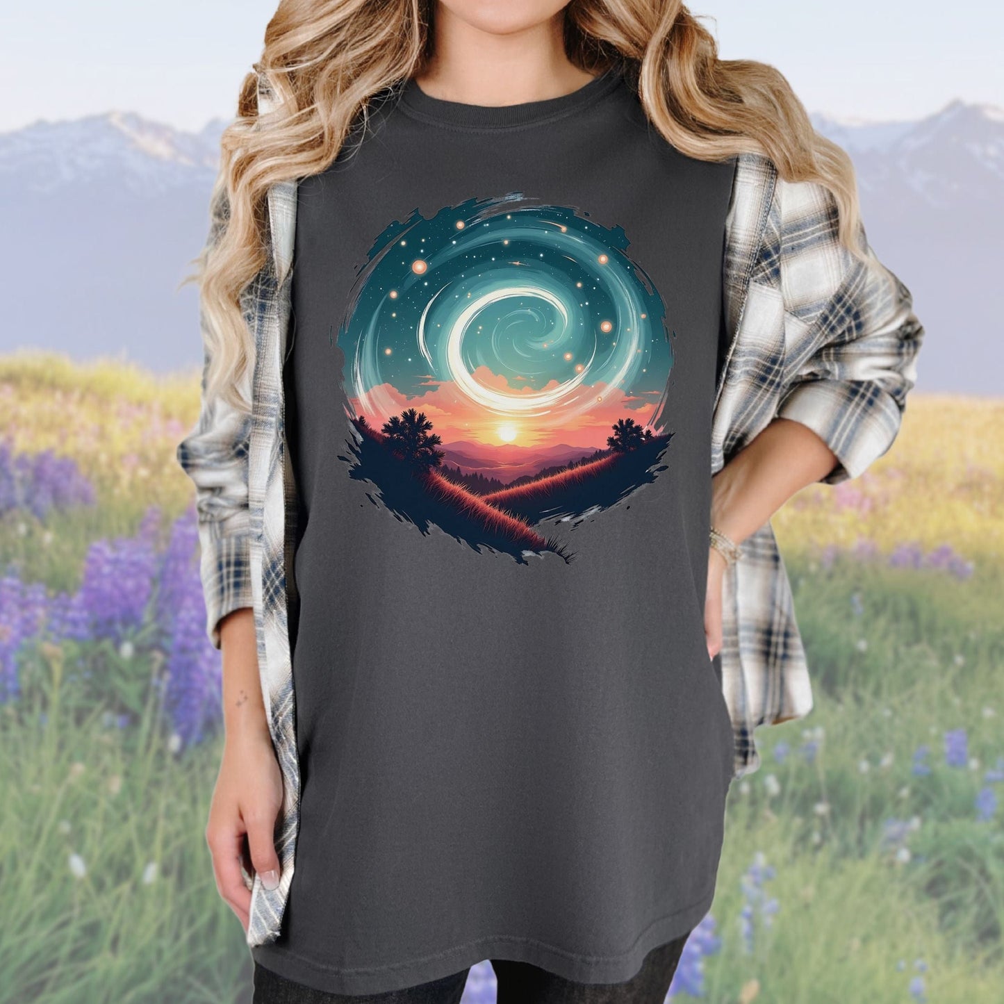 A person wearing a vintage-style graphic tee in a graphite color with a swirl pattern in the design, standing in a field with mountains in the background.