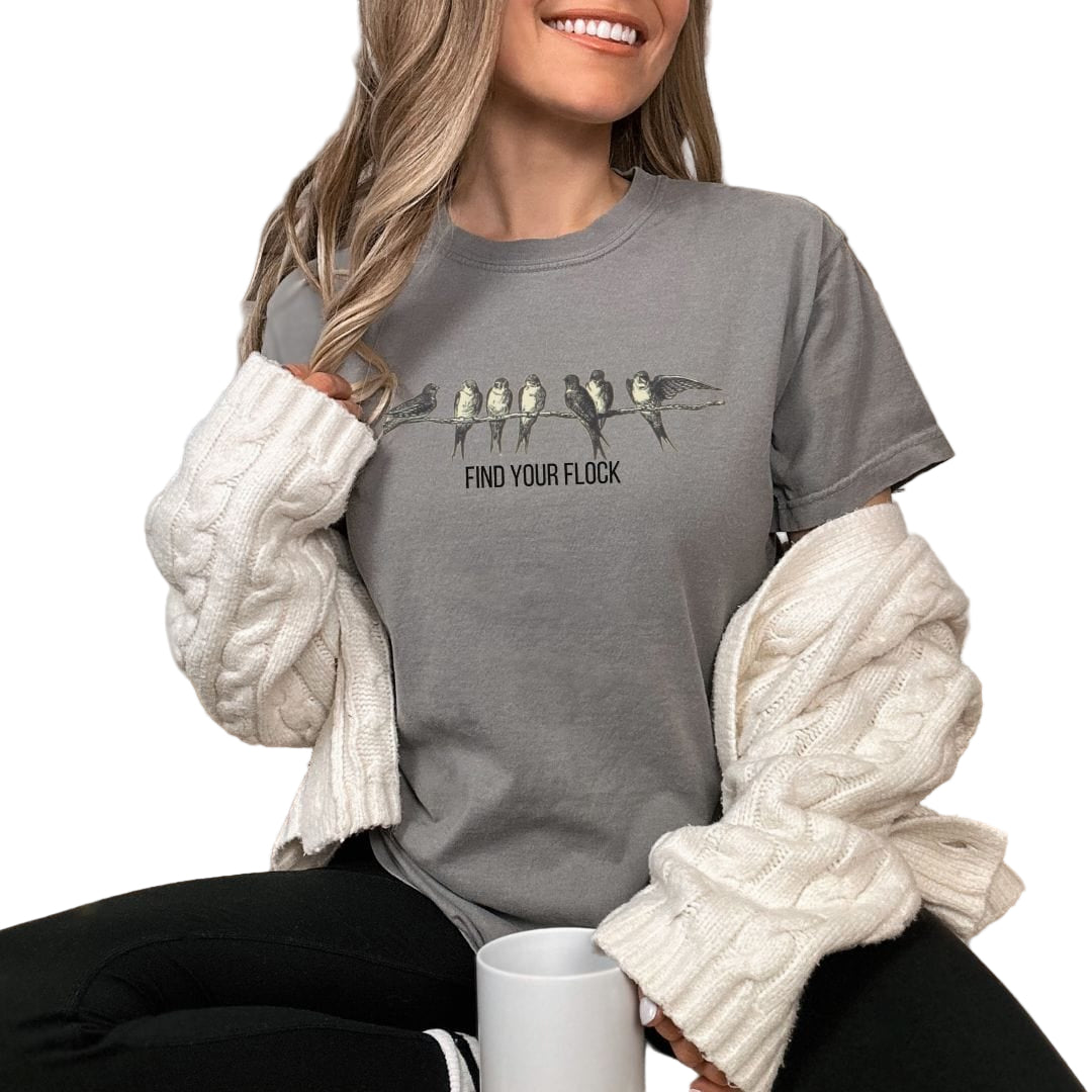 A person wearing a vintage-style gray t-shirt with bird illustrations and the text 'FIND YOUR FLOCK' on it, sitting in a cozy room with a knit blanket and a cup of coffee.