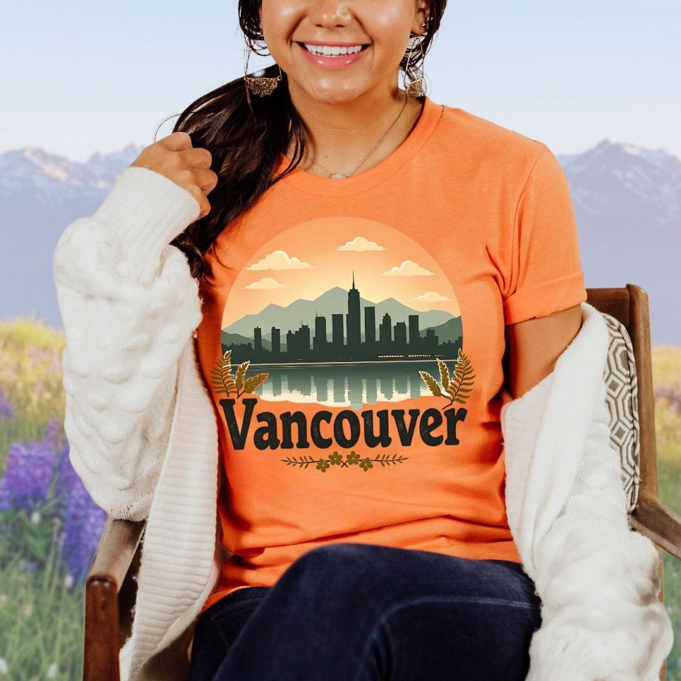 A person wearing an orange t-shirt with a cityscape design and the text 'VANCOUVER' on it, sitting in a field with mountains in the background.